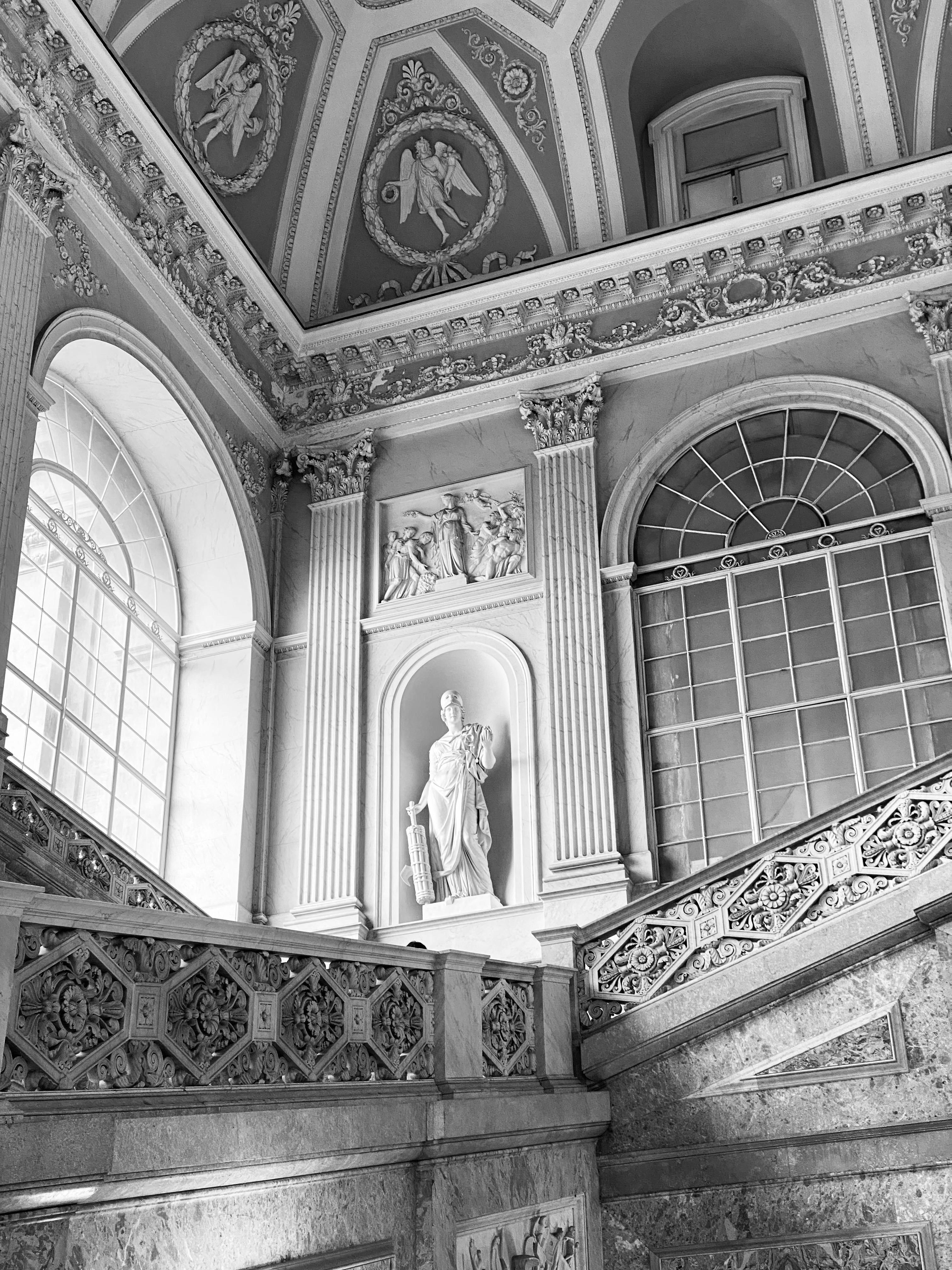 Black-and-white photograph of a monumental marble staircase with a classical statue in a niche beneath an ornate vaulted ceiling