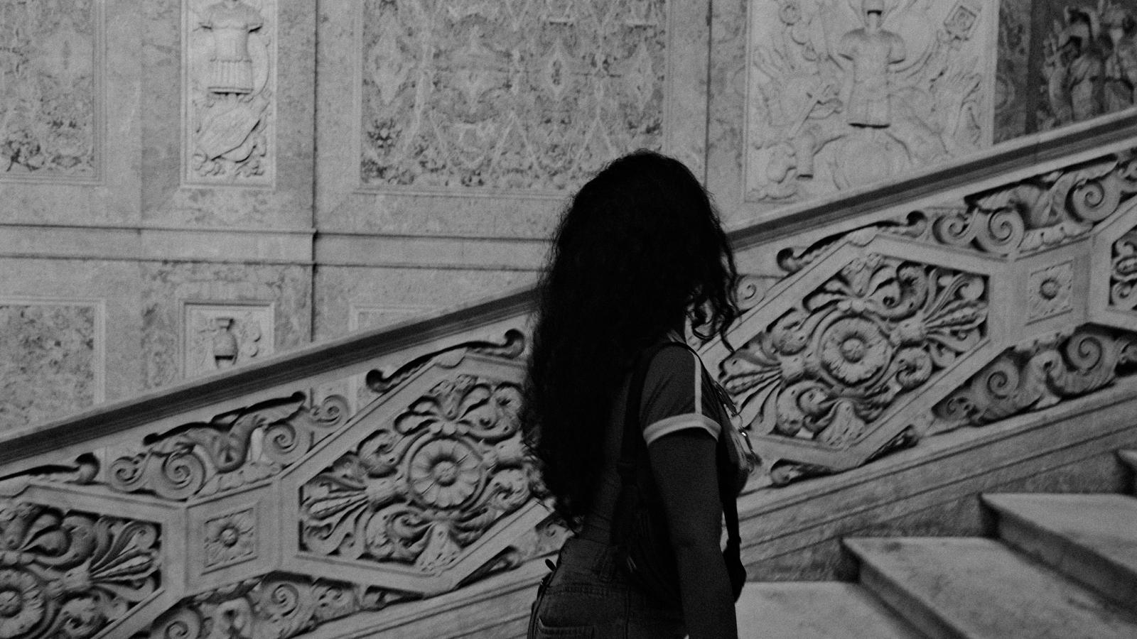 Black-and-white photograph of a woman with long dark curls seen from behind, walking beside a carved marble staircase balustrade