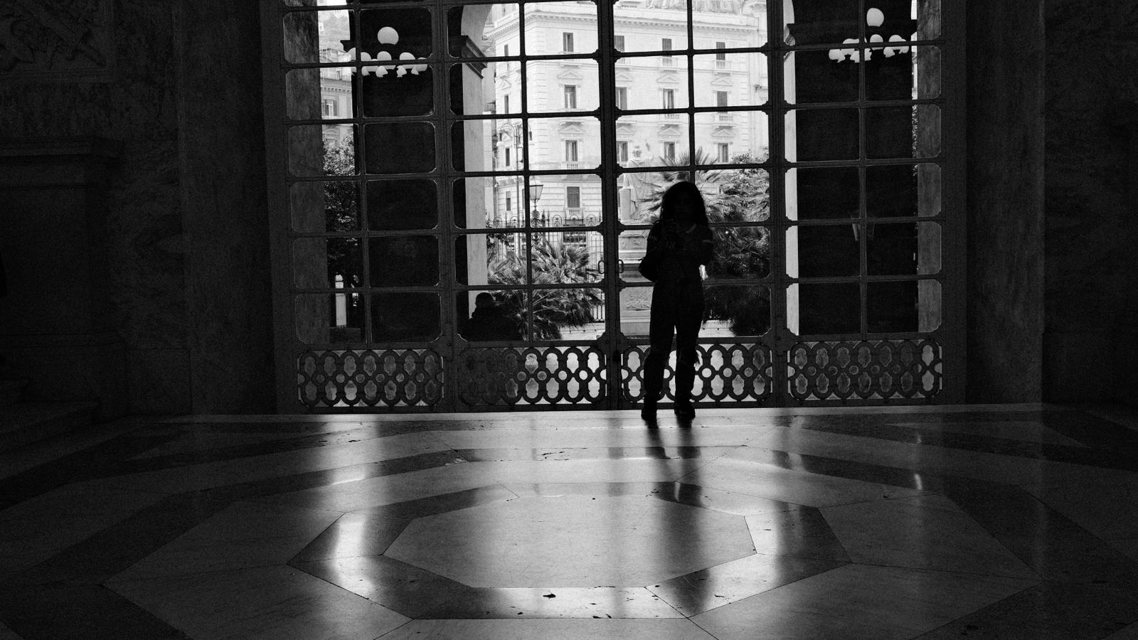 Black-and-white photograph of a silhouetted figure standing before a vast iron-framed window, light reflecting across a geometric marble floor