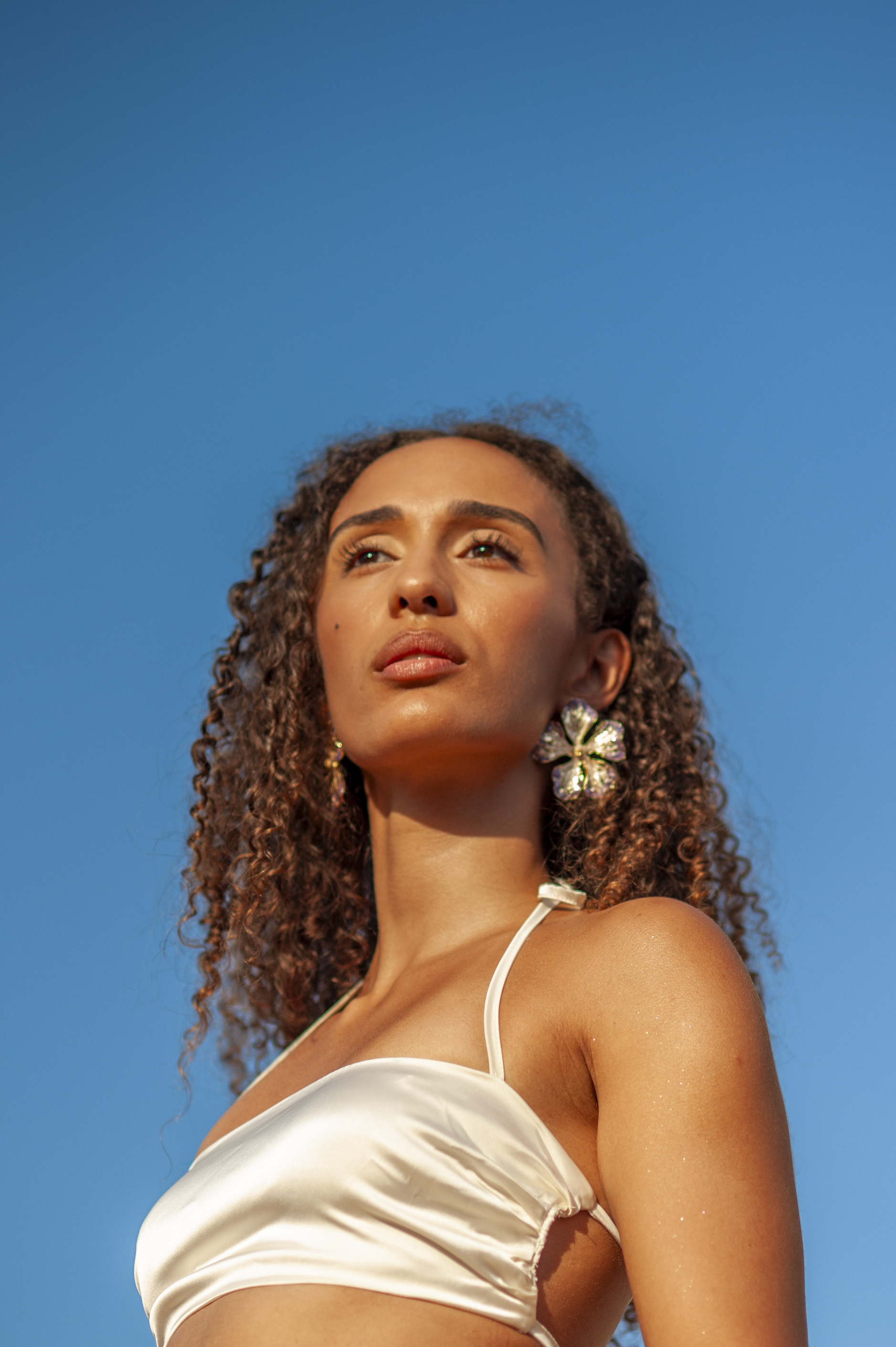 Low-angle portrait against a clear Mediterranean blue sky, model with long curls wearing a sculptural flower earring and ivory satin top