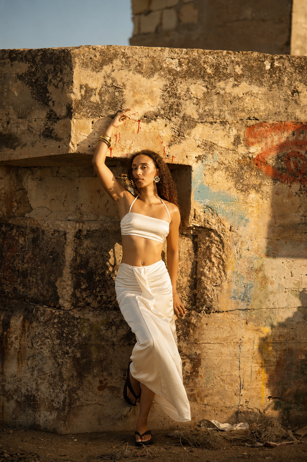 Model in an ivory satin top and skirt leaning against a weathered limestone wall with painted graffiti, gold bangles on a raised arm