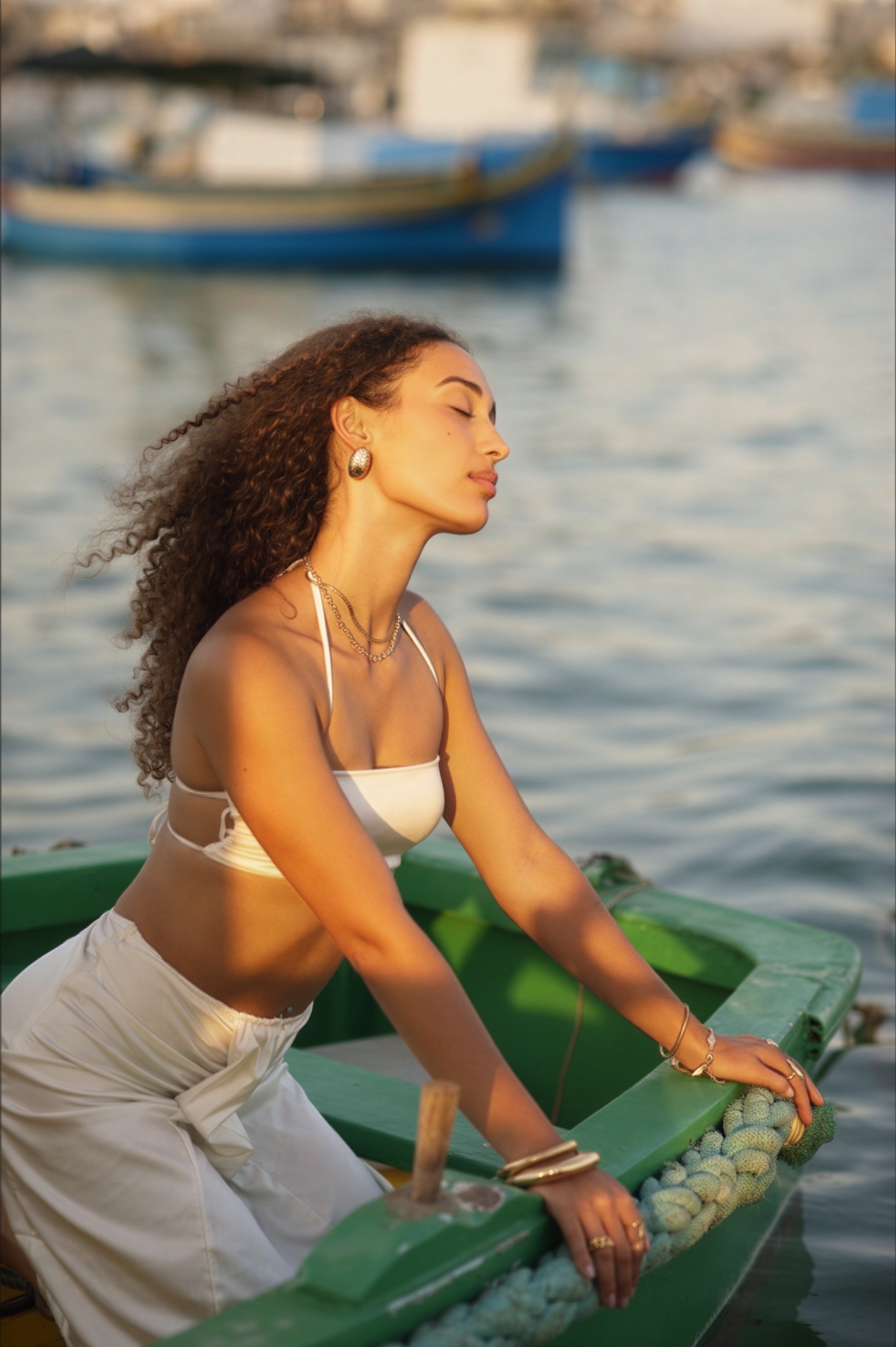 Model leaning over the edge of a green boat, eyes closed and head tilted to the sun, with blue luzzu boats blurred in Marsaxlokk harbour behind