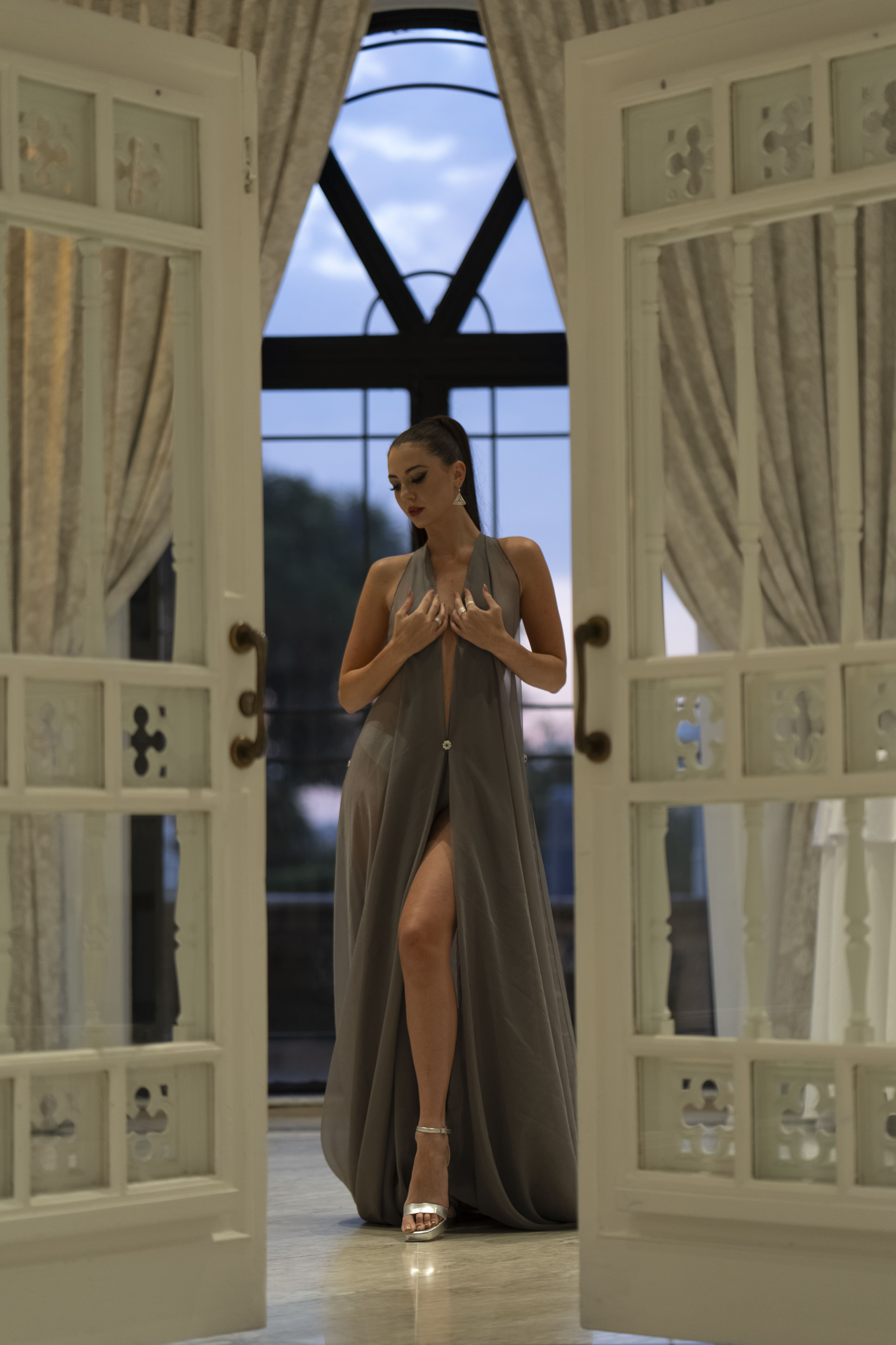 Model in a flowing grey chiffon gown, head lowered and hands at her chest, framed by carved white doors, damask curtains and a dusk sky through the window behind