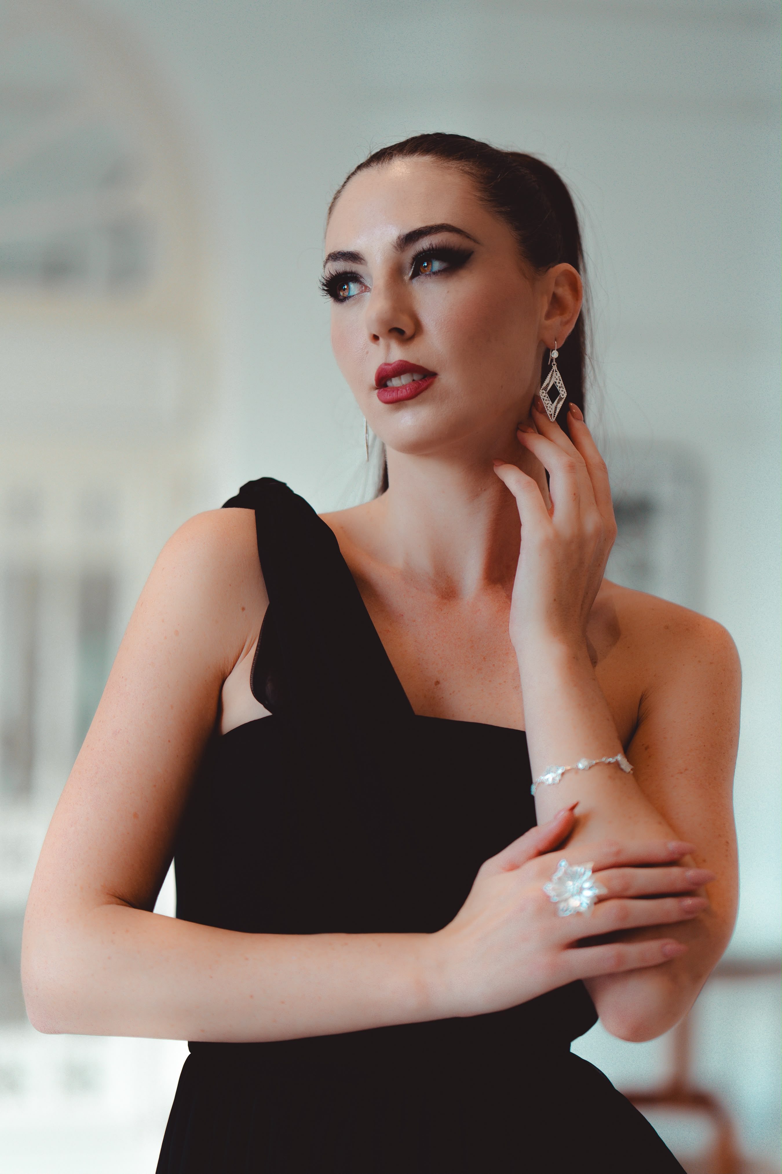 Close portrait of the model in a black one-shoulder dress, wearing silver filigree drop earrings, a delicate bracelet and a sculptural flower ring
