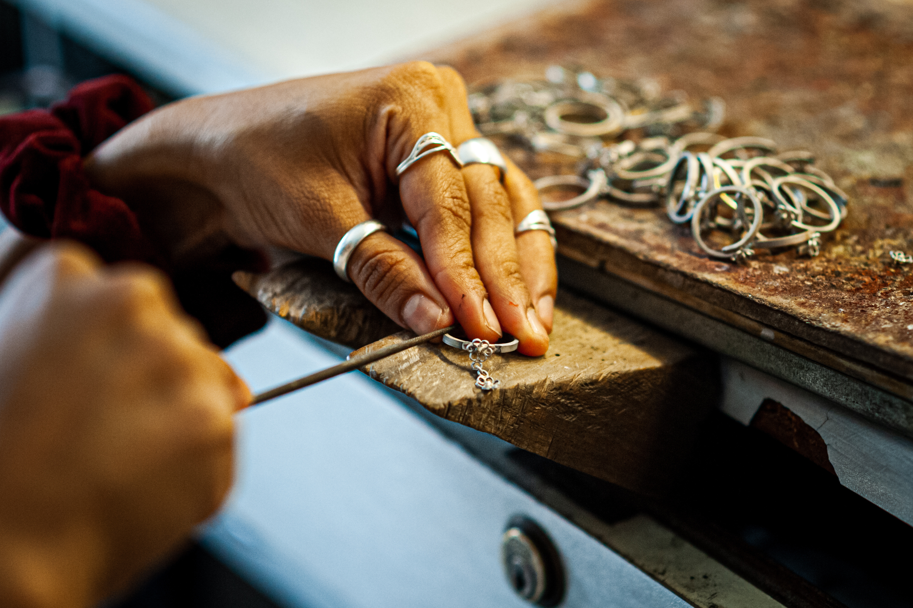 A silversmith's hands filing a filigree ring at a worn wooden bench, finished rings piled beside