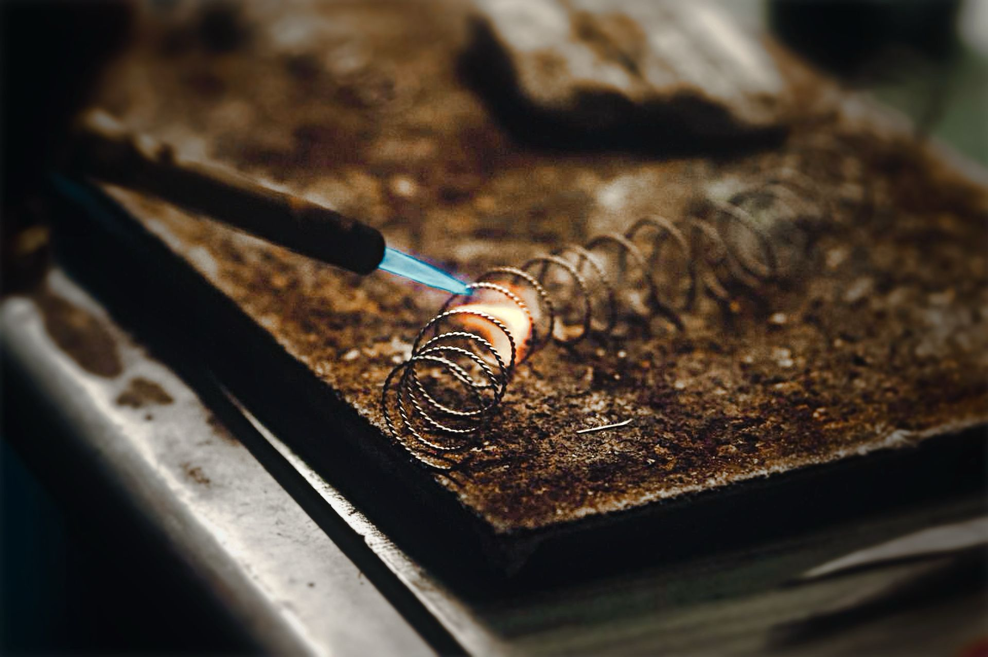 A torch flame heating coils of twisted silver wire on a worn workshop bench