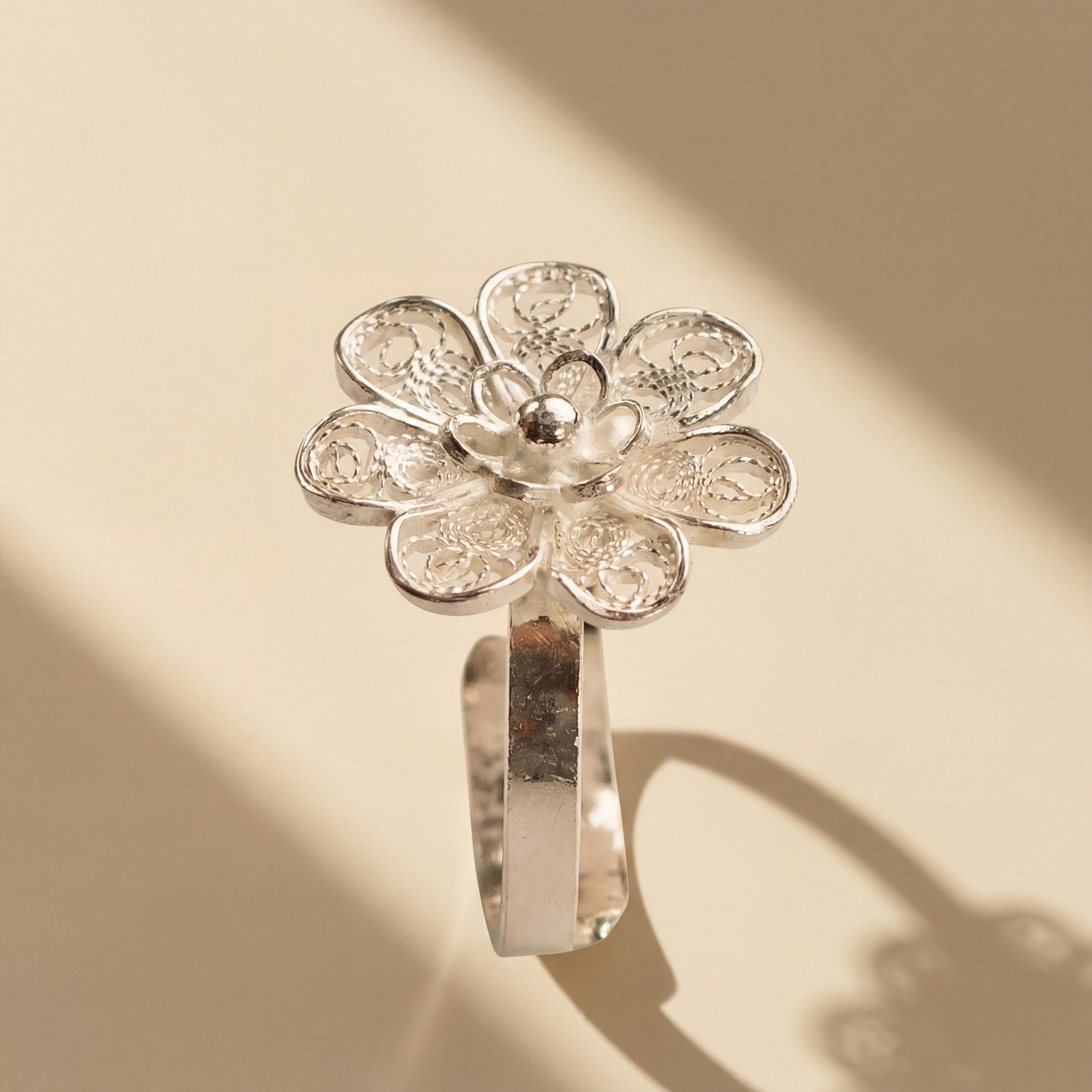 Filigree flower ring standing on a warm ivory surface with long diagonal shadows