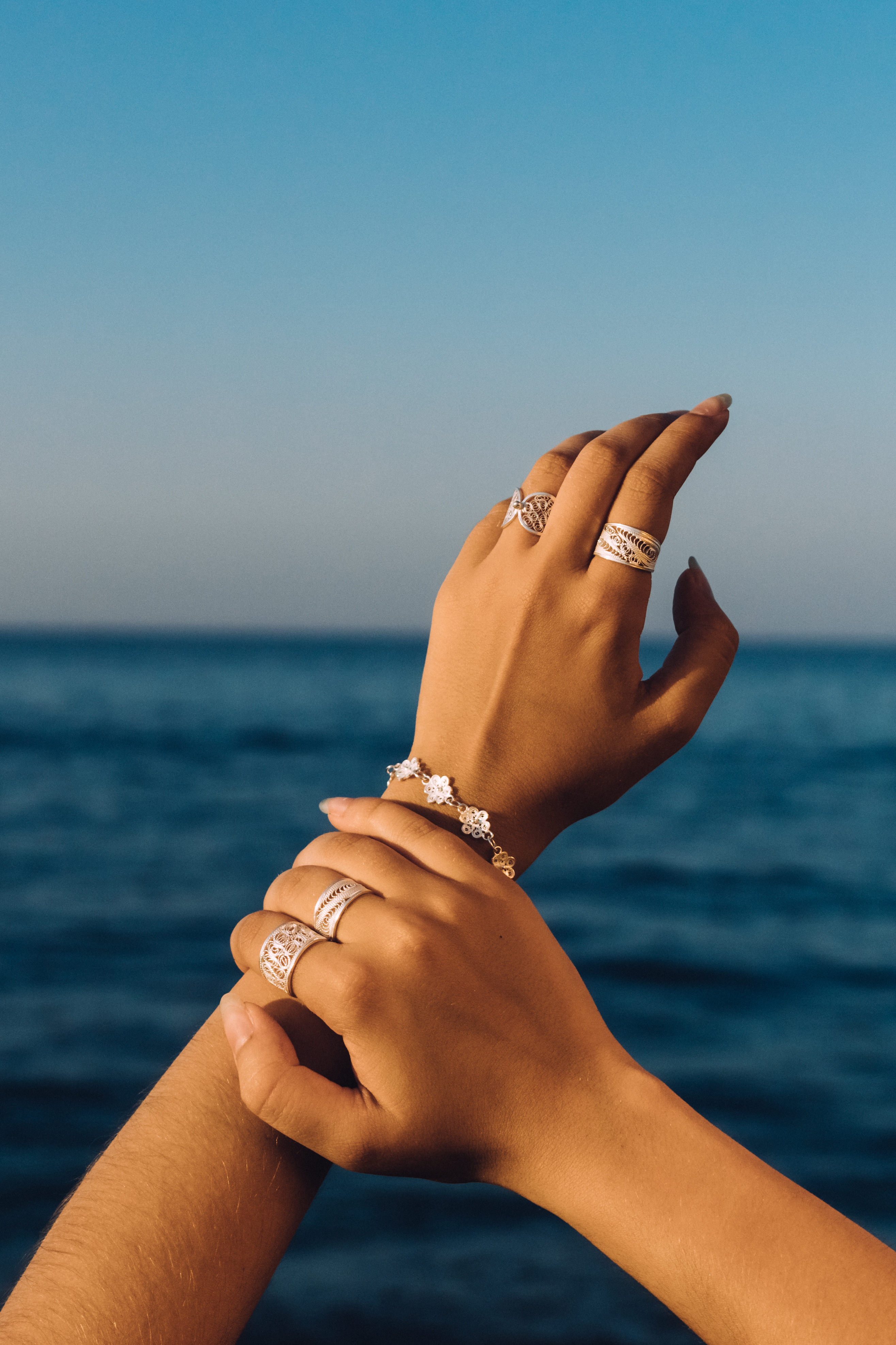 Hands with silver filigree rings and a flower bracelet raised against the Mediterranean sea