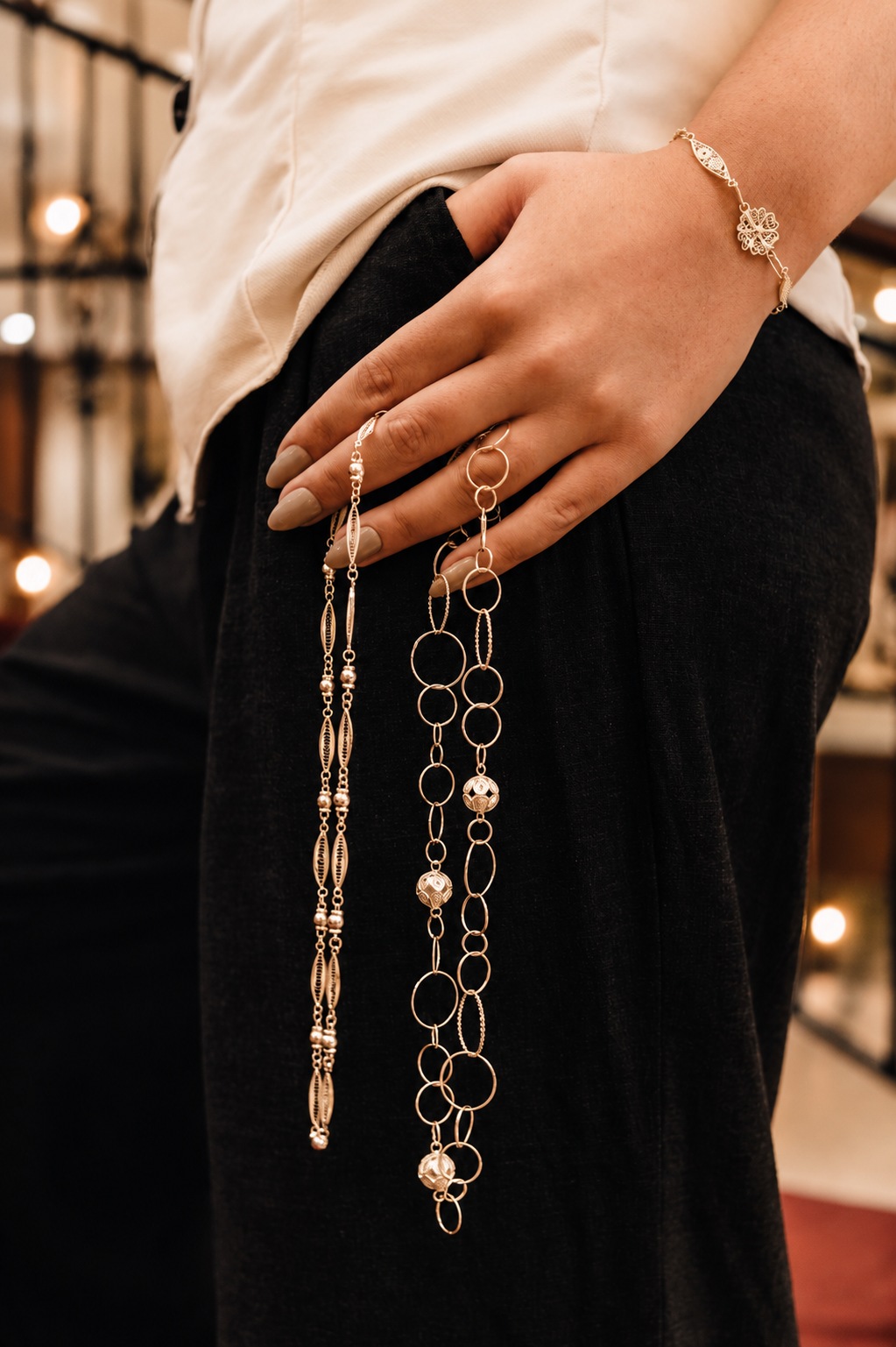 Hand at the hip holding long filigree chains in a warmly lit interior