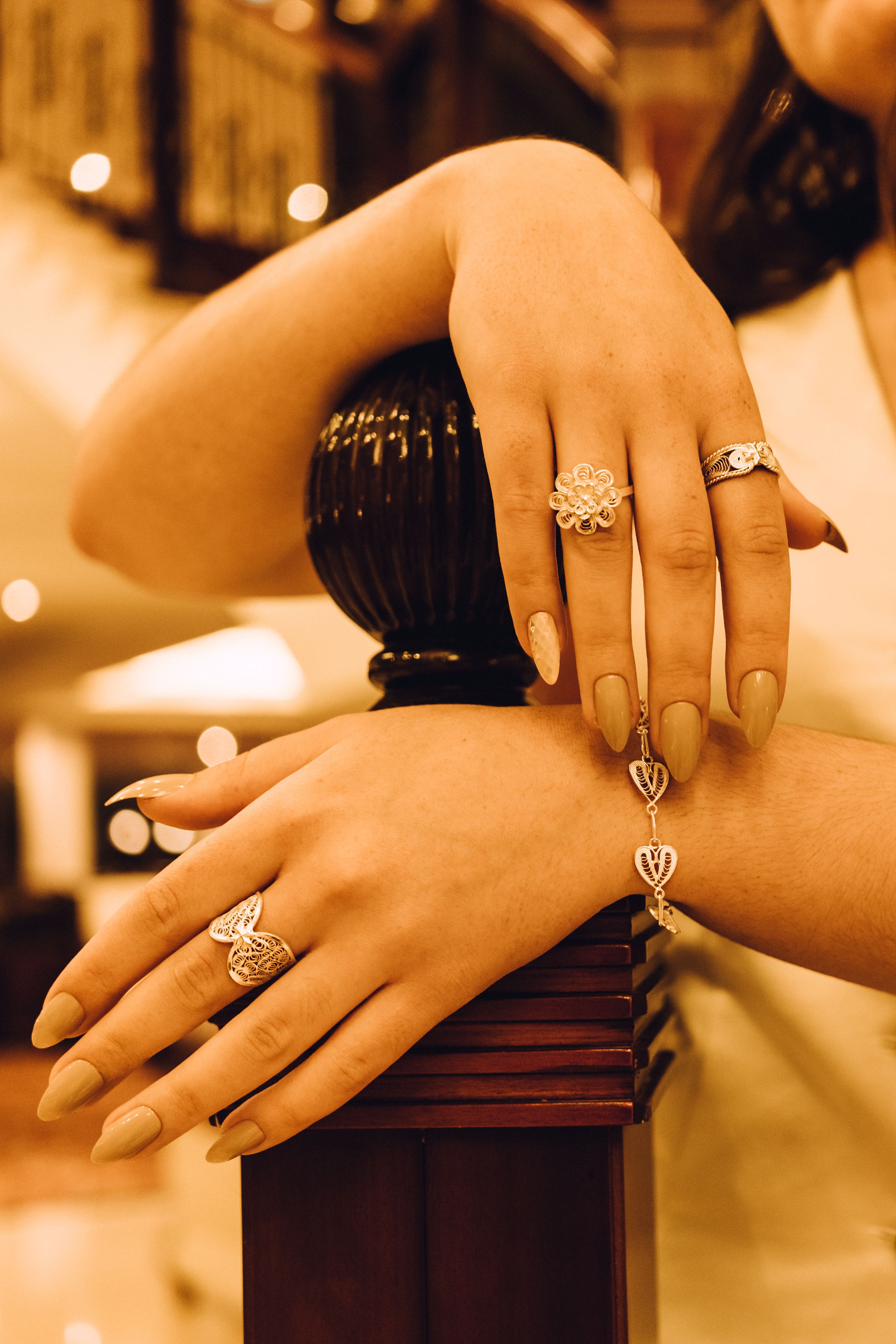 Crossed hands with filigree rings and a heart bracelet resting on a dark wooden newel post