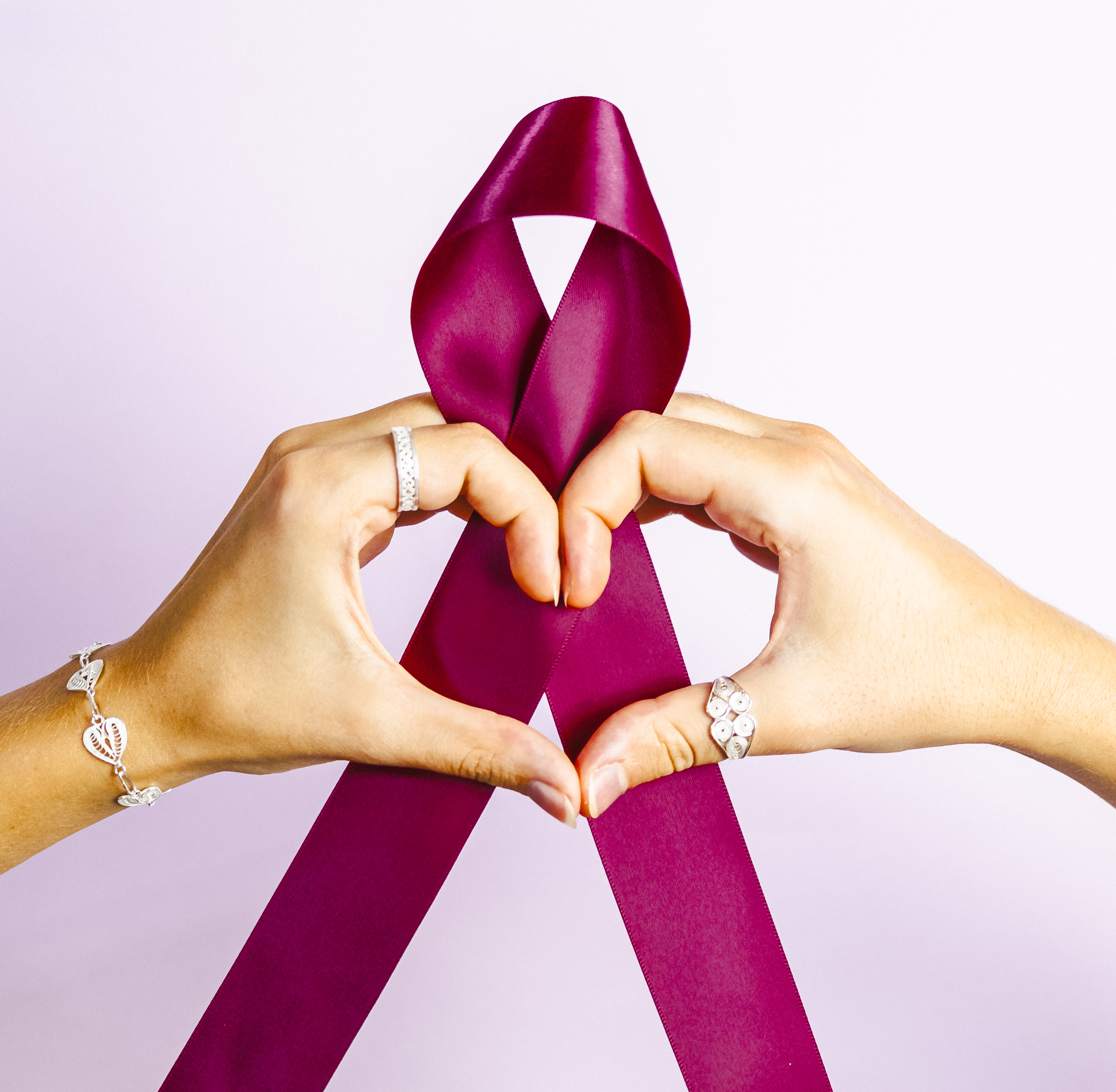 Two hands forming a heart around a burgundy ribbon, wearing filigree rings and a bracelet