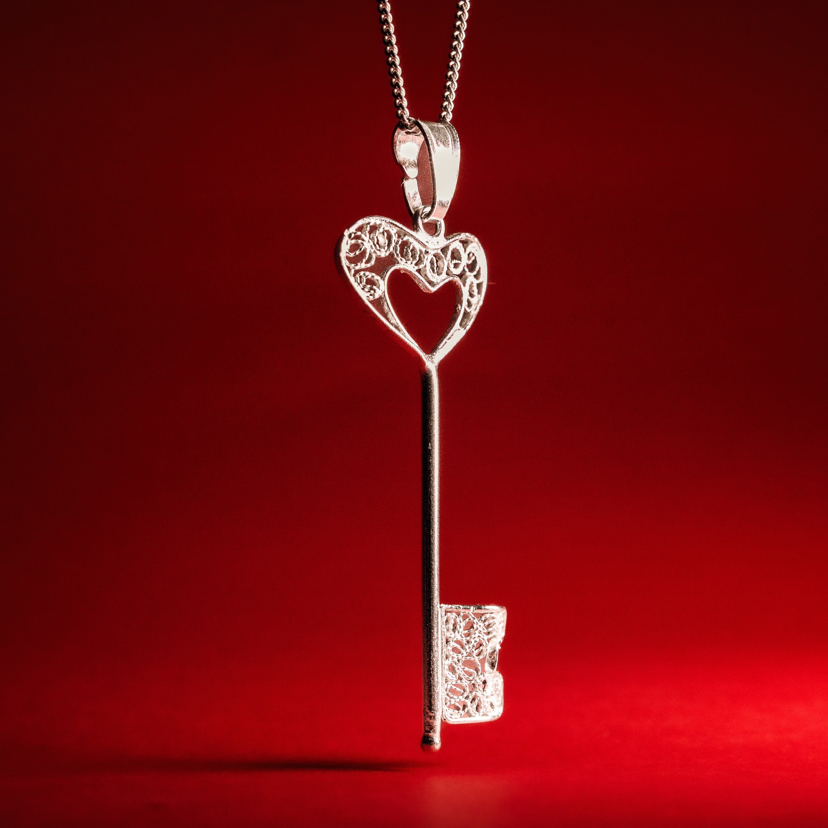 Silver filigree key pendant with a heart-shaped bow suspended against a deep red backdrop