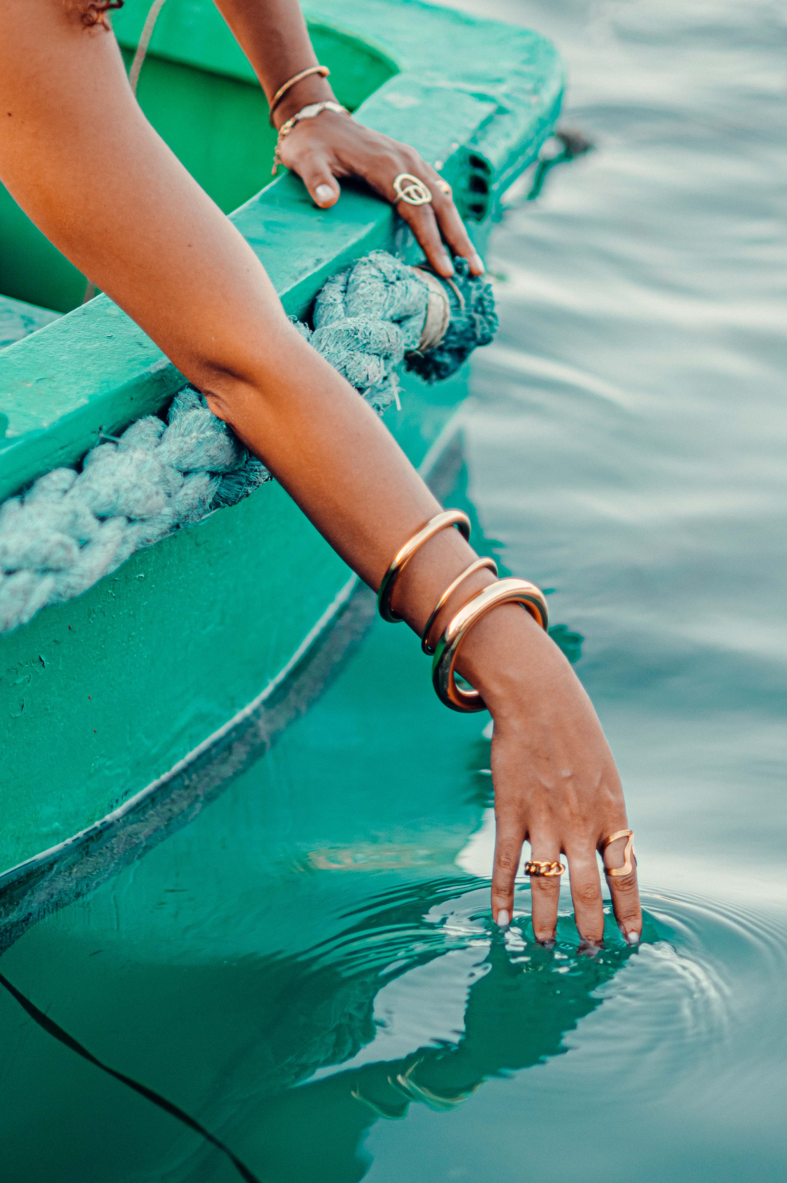 Woman’s arm with gold bangles and rings draped over a green boat, hand trailing in turquoise water